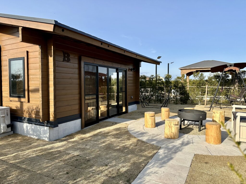 木製の外壁の建物。手前には丸太の椅子が配置されたファイヤーピットがあり、周辺には芝生の庭やブランコが見える