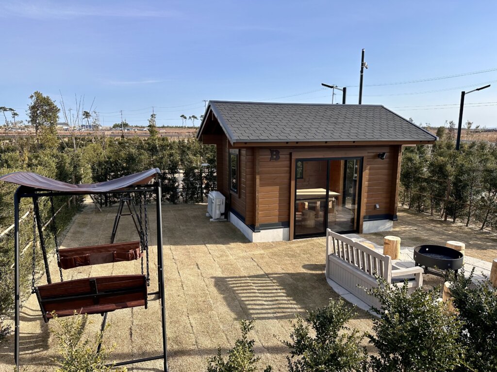 芝生の庭に設置された木製の小屋。手前にはブランコや丸太の椅子付きファイヤーピットが配置され、青空が広がる開放的な屋外空間