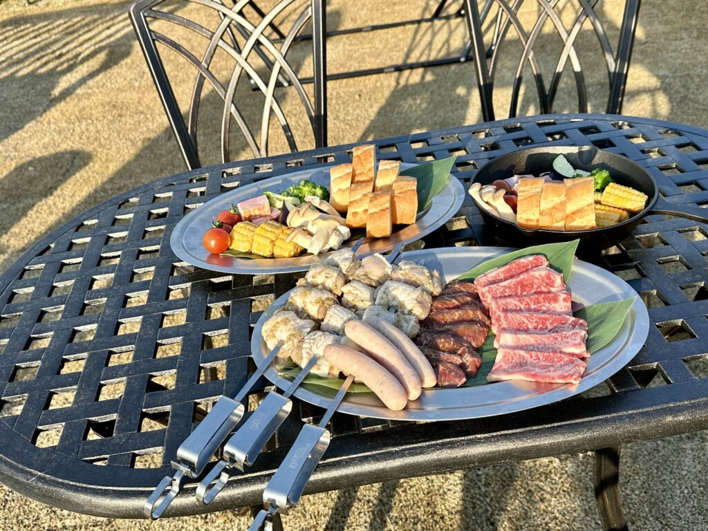 屋外の黒いメッシュ調テーブルの上に並べられた、牛肉や鶏肉の串刺し、ソーセージ、パン、野菜などが盛られたバーベキュー用の大皿とスキレット